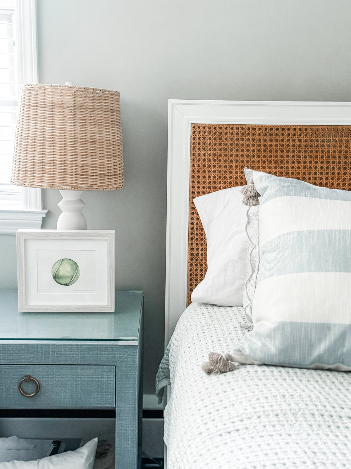 Bedroom with a bed featuring a wicker headboard, nightstand with lamp and framed picture, and light gray walls.